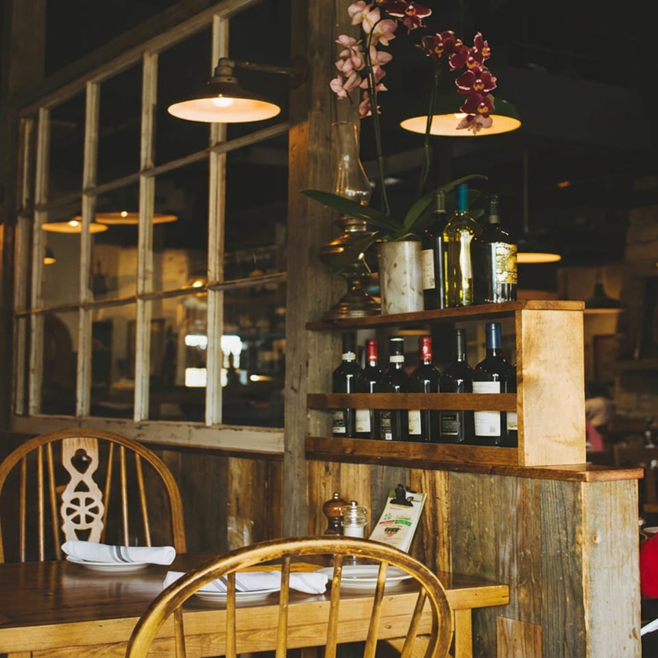 Cozy rustic restaurant interior with wooden tables, wine bottles, hanging lights, orchids, and a diner in a red jacket.
