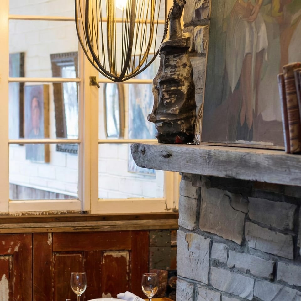 Rustic restaurant interior with a stone fireplace, wooden mantel, vintage books, sculptural decor, and dining table by sunlit windows.