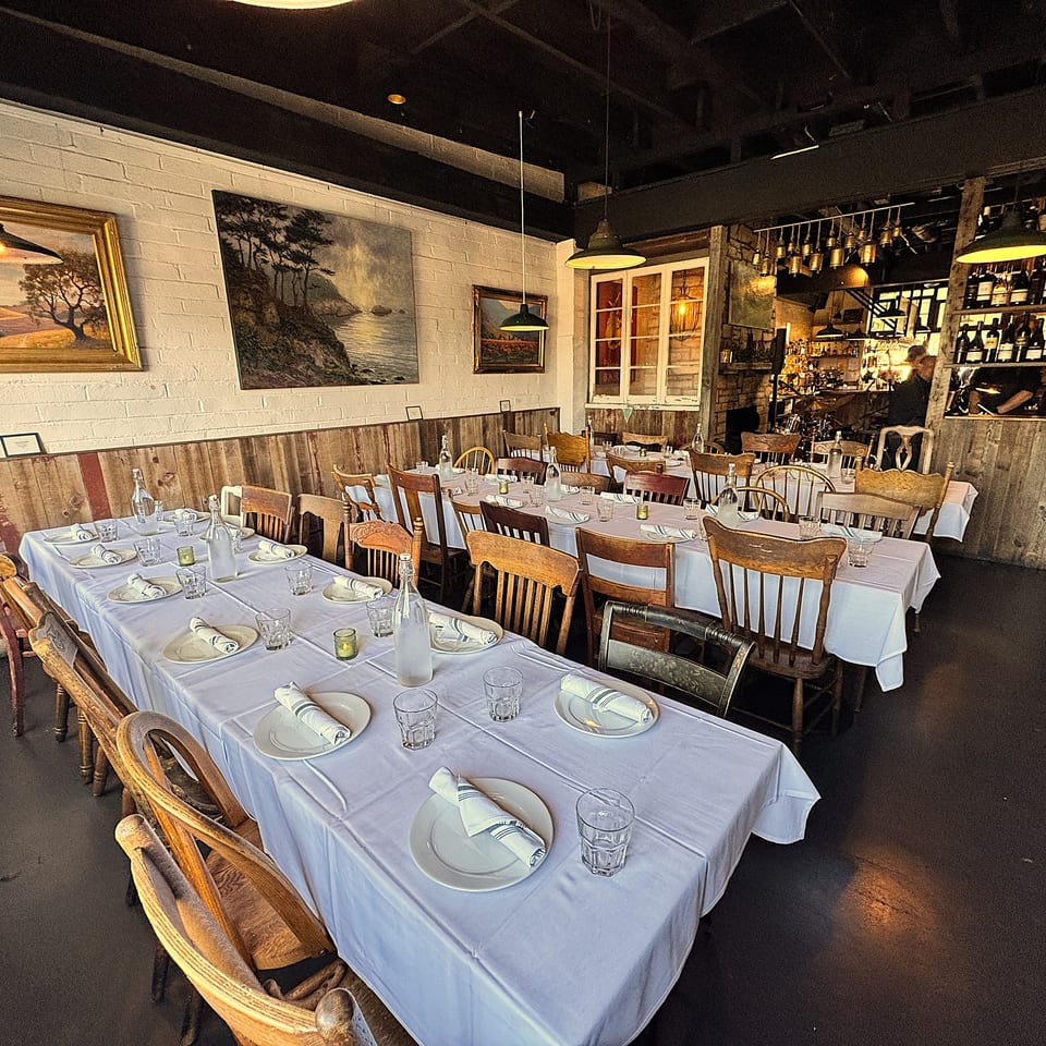Warmly lit restaurant dining room with white tablecloths, set tables, rustic artwork, wooden chairs, and a stocked bar.