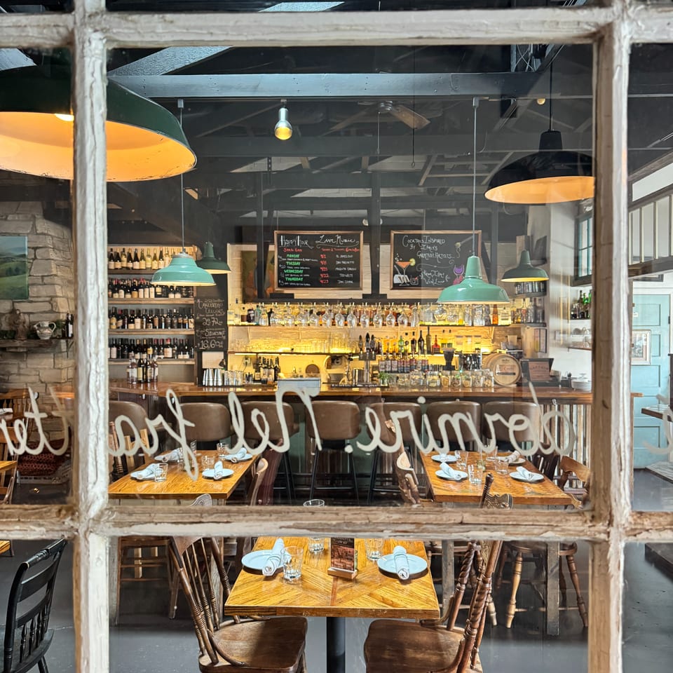 Rustic restaurant interior with a fully stocked bar, chalkboard menus, pendant lights, and neatly set wooden dining tables.