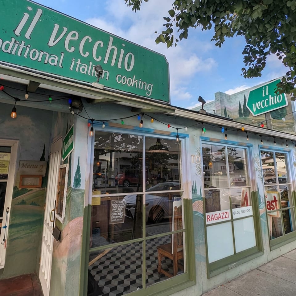 Exterior of Il Vecchio traditional Italian restaurant with colorful string lights, large windows, tree-shaded sidewalk, and an open sign.