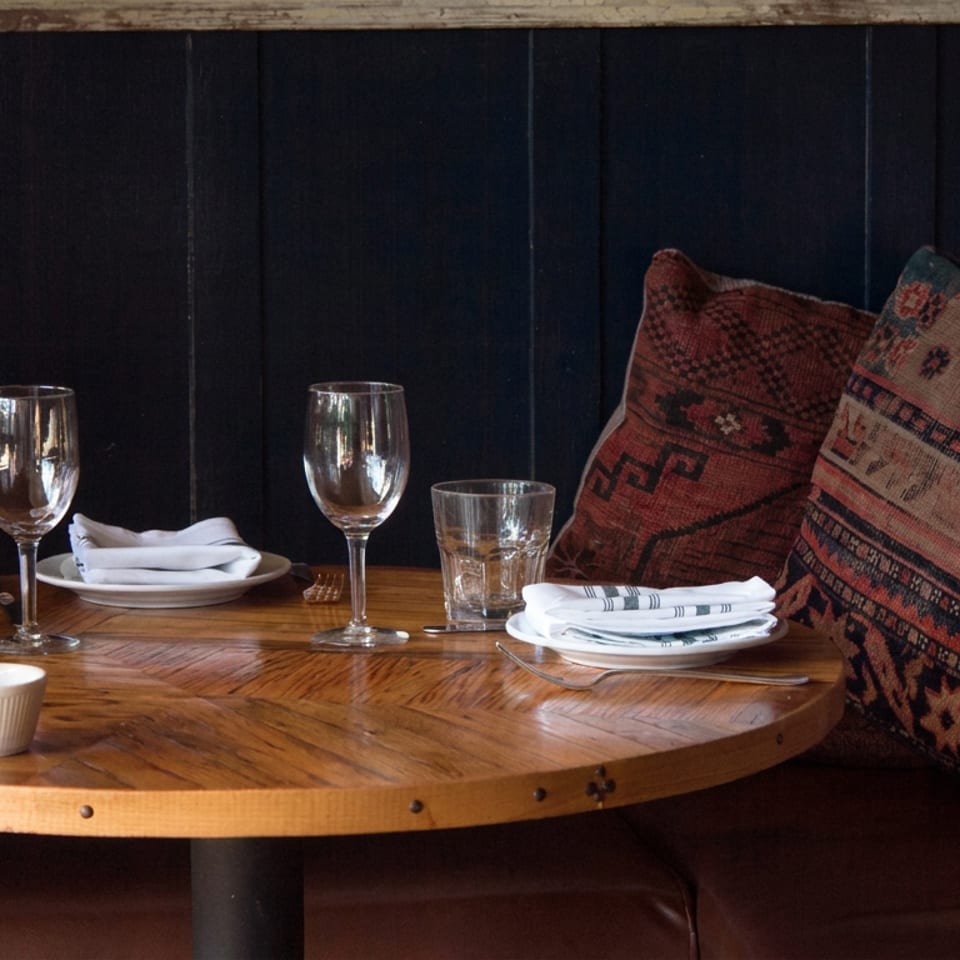 Cozy restaurant booth with a round wooden table set with wine glasses, water glasses, plates, folded napkins, and patterned pillows.