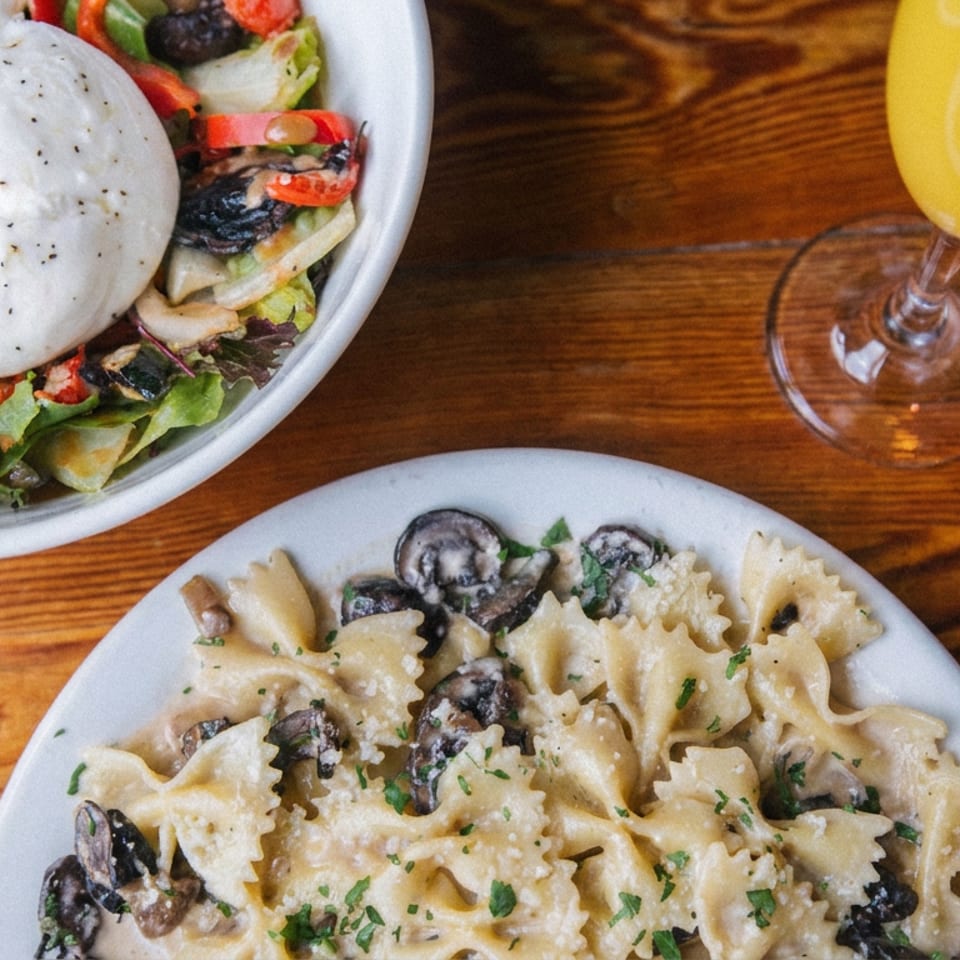 Overhead view of creamy mushroom farfalle, a burrata-topped vegetable salad, and a yellow drink on a wooden restaurant table.